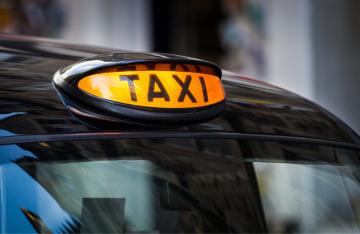 Photograph of an illuminated orange Taxi sign on the roof of a black cab with blurred streets in the background.